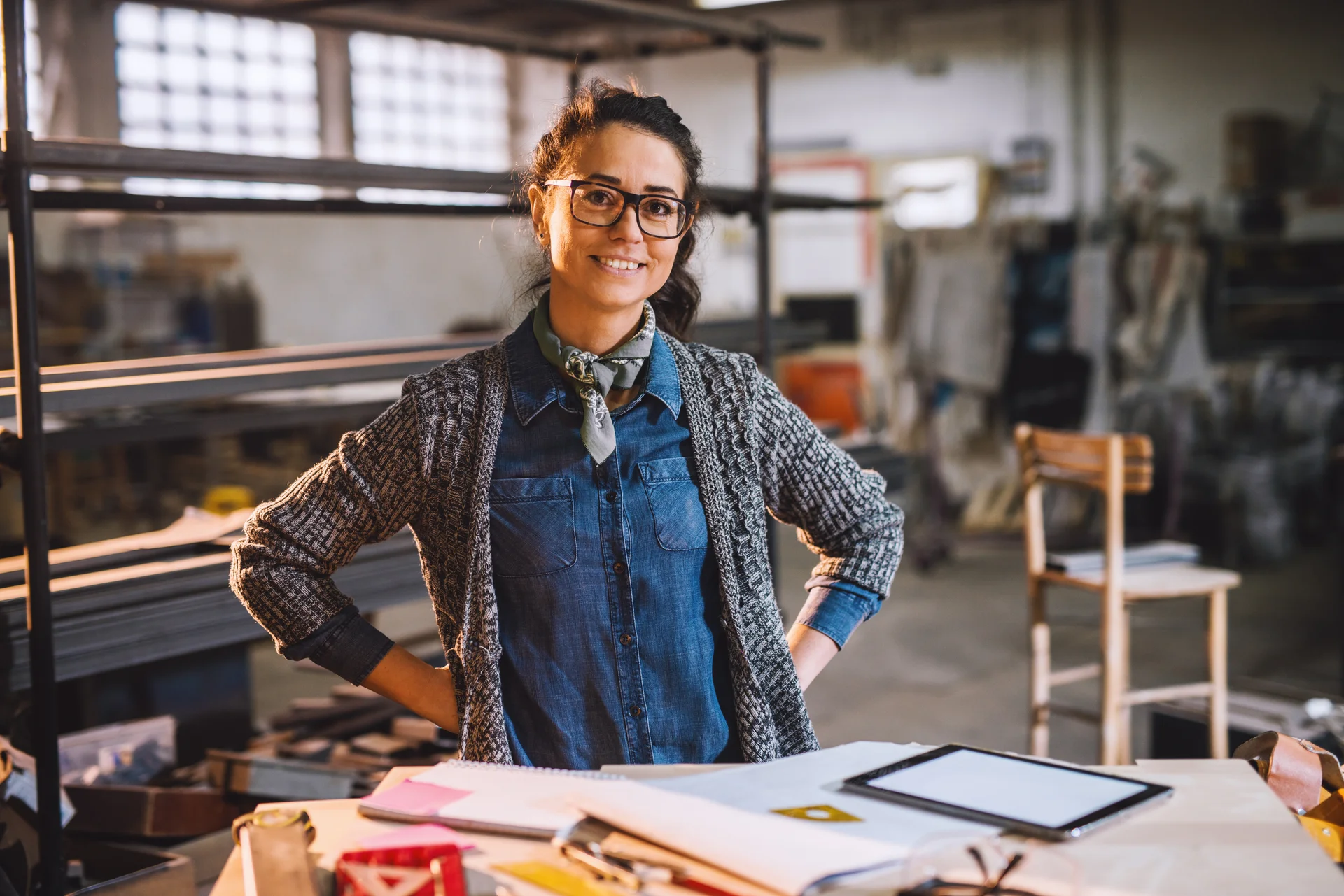 Eine Frau mit Brille und einem grauen Strickpullover, die in einer Werkstatt an einem Tisch steht. Sie lächelt und ist von verschiedenen Arbeitsmaterialien umgeben, darunter Papiere und ein Tablet.