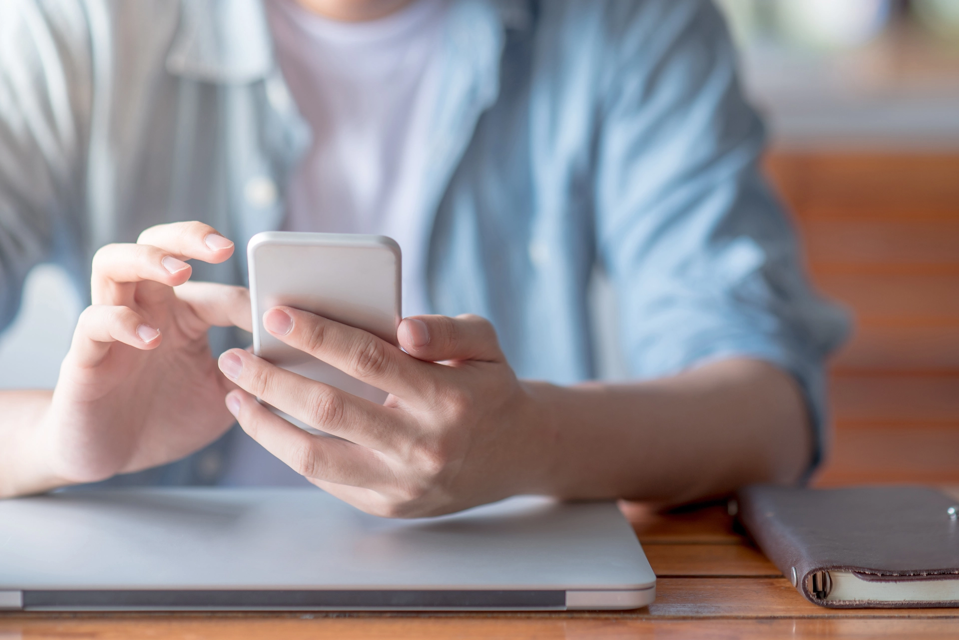 Ein Mann hält ein Smartphone in der Hand, während er auf einem Tisch mit einem Laptop arbeitet. Die Szene ist im Vordergrund fokussiert, und der Mann trägt ein Hemd mit kurzen Ärmeln.