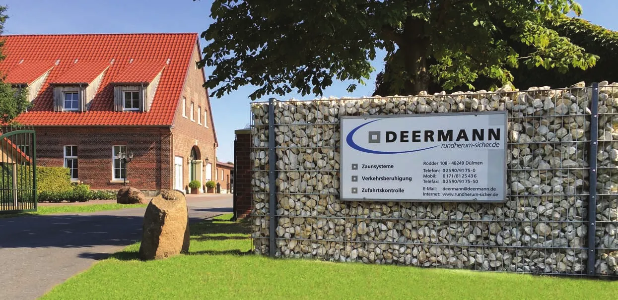 Das Bild zeigt ein Gebäude mit einem rot gedeckten Dach, vermutlich ein Wohn- oder Geschäftsgebäude, das von viel Grün umgeben ist. Vor dem Gebäude befindet sich eine Steinmauer oder ein Zaun mit einem Schild, auf dem „DEERMANN“ steht, zusammen mit Kontaktinformationen und Dienstleistungen im Bereich Zaunbau und Sicherheit. Die Umgebung wirkt gepflegt und sonnig, was auf eine angenehme Atmosphäre hinweist.
