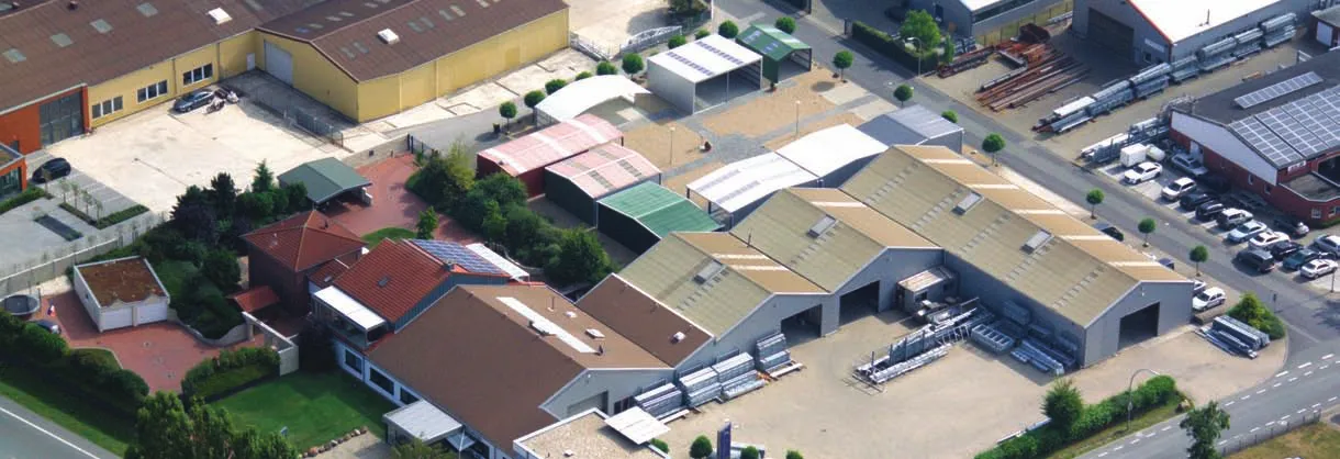 Das Bild zeigt eine Luftaufnahme eines Industriegebiets. Es sind mehrere Gebäude mit unterschiedlich gefärbten Dächern zu sehen, darunter grüne, rote und beige Dächer. Die Anordnung umfasst Freiflächen, die möglicherweise als Park- oder Lagerflächen genutzt werden, und rund um die Gebäude befindet sich viel Grün und Bäume. Einige der Bauwerke scheinen Lagerhallen oder Werkstätten zu sein, und auf dem Gelände sind Fahrzeuge geparkt. Insgesamt wirkt die Anlage wie ein gewerbliches oder industrielles Areal.