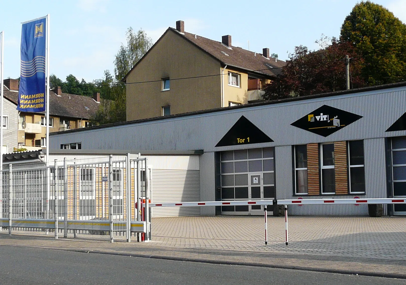 Das Bild zeigt ein Gebäude mit modernem Design und einem großen Eingang, der mit „Tor 1“ gekennzeichnet ist. Vor dem Gebäude befindet sich ein eingezäunter Bereich, und die Fassade besteht aus Metall mit großen Fenstern. Im Hintergrund sind Wohngebäude und Bäume zu sehen. Außerdem ist ein Fahnenmast mit einer blauen Fahne sichtbar. Insgesamt wirkt die Umgebung wie ein Industrie- oder Gewerbegebiet.