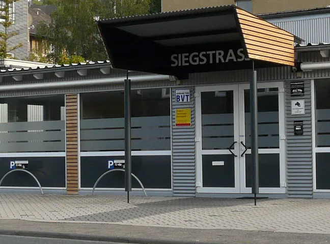 Das Bild zeigt einen Gebäudeeingang mit einem Schild, auf dem „SIEGSTRAS“ steht. Die Architektur ist modern, mit einem Dach, das Holzakzente aufweist, und Glastüren. Es sind auch Parkschilder zu sehen, die auf ausgewiesene Parkbereiche hinweisen. Die Umgebung ist gepflastert, und im Hintergrund sind einige Bäume zu erkennen.